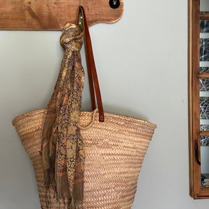 Puede incluir: Un gran bolso tote de paja tejida con asas de cuero marrón cuelga de un gancho de pared de madera. Una bufanda estampada en tonos dorados y marrones está drapeada sobre el bolso. El bolso mide aproximadamente 46 cm de ancho.