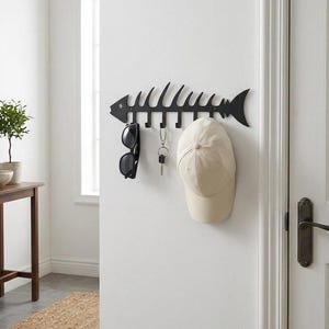 May include: A black metal wall-mounted hook rack shaped like a fish skeleton. The rack features five downward-facing hooks holding a pair of black sunglasses, a silver metal keyring with keys, and a beige baseball cap against a white wall.