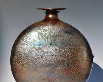 May include: A round, metallic vase with a wide, flat base and a narrow neck. The vase has a textured, iridescent finish with shades of copper, bronze, and silver.