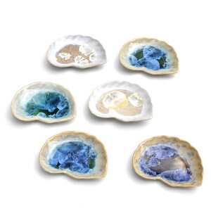 May include: A collection of six small, shell-shaped ceramic dishes featuring crystalline glaze patterns. Each dish displays unique, organic radial formations in shades of blue, white, and beige, with scalloped edges and a smooth, glossy finish.