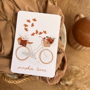 Peut inclure: Une carte blanche avec une illustration de vélo, avec des citrouilles et des fleurs dans les paniers, et des feuilles d'automne orange. Le texte "pumpkin lover" est écrit en cursive. La carte est placée dans un panier en osier avec un pull brun.