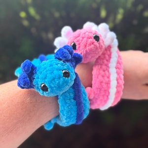 May include: Two plush axolotl toys are shown. One is blue with dark blue stripes, and the other is pink with white accents. Both have black eyes and are made of a soft, textured material. The toys are designed to be worn as bracelets.
