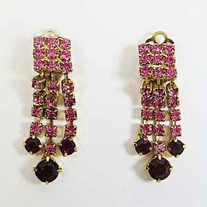 May include: A pair of dangle earrings with pink and red crystal stones. The earrings have a gold tone finish.