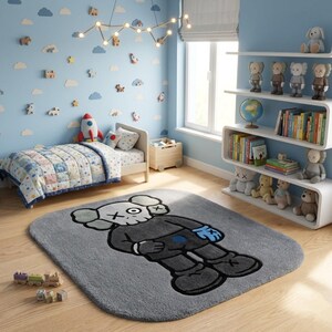 K&ouml;nnte beinhalten: Ein grauer rechteckiger Teppich mit einer Cartoonfigur in einem schwarzen Anzug, wei&szlig;em Kopf und durchgestrichenen Augen. Der Teppich befindet sich in einem Kinderzimmer mit Bett, Regalen mit Spielzeug und einer blauen Wand mit Wolken- und Raketendekorationen.