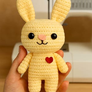 Könnte beinhalten: Ein handgehäkelter, gelber Hase mit schwarzen Augen, einer rosa Nase und einem roten Herz. Der Hase hat lange Ohren und wird in einer Hand gehalten. Im unscharfen Hintergrund ist eine Nähmaschine zu sehen.