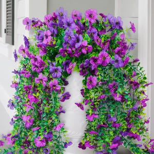 Könnte beinhalten: Eine lebendige Präsentation künstlicher Blumen in Lila- und Magentatönen. Die kaskadenartige Anordnung zeigt üppiges grünes Laub und Blüten und erzeugt ein volles und strukturiertes Aussehen. Die Blumen sind in einem weißen Pflanzgefäß angeordnet, ideal für einen Farbtupfer.