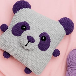 May include: A handmade, square-shaped panda pillow in gray and purple. The pillow features a panda face with purple eyes, ears, and accents. A ball of purple yarn sits nearby on a pink fabric background.