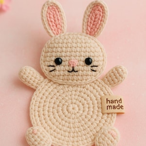 May include: A crocheted bunny coaster in beige with pink accents on the ears, nose, and feet. The bunny has black eyes and whiskers. A small tag that says "hand made" is attached to the coaster.
