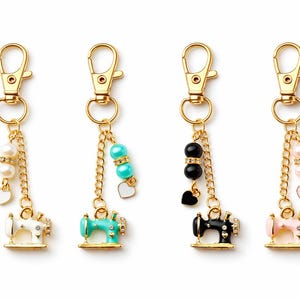 May include: Four gold keyrings with sewing machine charms. Each charm is a different colour: white, turquoise, black, and pink. Each keyring has a gold chain with beads and a heart charm.