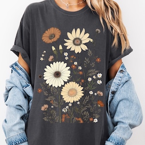May include: Dark gray t-shirt featuring a floral design with daisies and other wildflowers in shades of brown, cream, and orange. The flowers are set against a backdrop of green leaves and stems. The shirt has rolled-up sleeves.