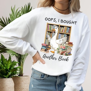 Puede incluir: Sudadera blanca con un diseño de un ganso frente a una estantería y pilas de libros. El texto dice "OOPS, I BOUGHT Another Book" en letras negras. El ganso es blanco con patas y pico amarillos.