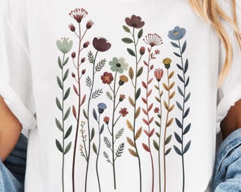 May include: White t-shirt featuring a colorful floral design. The graphic includes various wildflowers in shades of red, blue, green, and yellow. The flowers are arranged on slender stems, creating a vertical pattern.