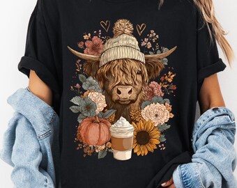 May include: Black t-shirt featuring a Highland cow wearing a knit hat, surrounded by flowers, a pumpkin, a sunflower, and a coffee cup. The design has a fall theme with warm colors and a whimsical style.