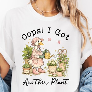 May include: White t-shirt with a whimsical graphic of a duck wearing a pink apron and hat, watering plants. The text "Oops! I Got Another Plant" is printed below the illustration. The design features potted plants and butterflies.
