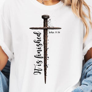May include: White t-shirt featuring a graphic of a rusty nail in the shape of a cross. The text "It is finished" is vertically aligned on the left side, with "John 19:30" on the right. The design has a distressed, vintage look.