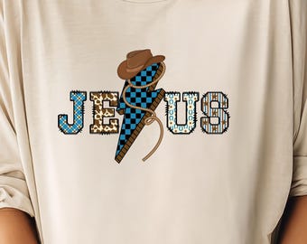 Könnte beinhalten: Beiges T-Shirt mit dem Wort "JESUS" in einem Western-Design. Das "J" und das "S" sind blau, das "E" ist braun und das "U" ist mit Hufeisen verziert. Ein brauner Cowboyhut und ein blauer und schwarzer Blitz befinden sich in der Mitte.