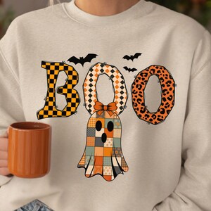 Op de afbeelding: Een lichtbeige sweatshirt met het woord "BOO" in een Halloween-ontwerp. De "B" is oranje en zwart geblokt, de eerste "O" is oranje en wit geblokt en de tweede "O" is oranje met een zwarte luipaardprint. Een patchwork spook staat in het midden.