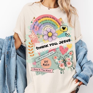 May include: Cream-colored t-shirt with a colorful graphic design. The design includes a rainbow, flower, butterfly, hearts, and text that reads "I have set my rainbow in the clouds," "thank you, Jesus," and "I AM A CHILD OF GOD."