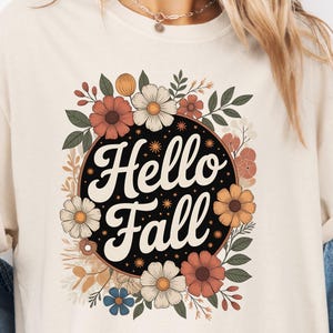 May include: Cream-colored sweatshirt featuring a floral design with the words "Hello Fall" in a circular black frame. The floral arrangement includes orange, white, and brown flowers with green leaves, creating a seasonal autumn theme.