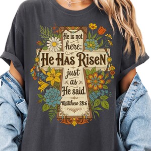May include: Dark gray t-shirt featuring a wooden cross design with floral accents and the text "He is not here; He Has Risen just as He said. Matthew 28:6". The design includes yellow, blue, and orange flowers.