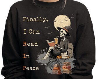 May include: Black sweatshirt with a Halloween-themed graphic. The design features a skeleton wearing a witch hat, reading a book, and drinking from a cup. The text reads "Finally, I Can Read In Peace."