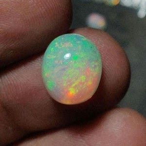 May include: An oval-shaped opal gemstone, displaying a play of color with green, orange, and white hues. The gemstone is held between fingers, showcasing its polished surface and internal iridescence. The opal's colors shift with light.