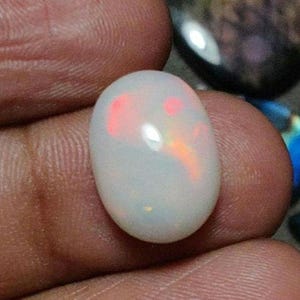 May include: An oval-shaped, white opal gemstone with internal flashes of red and orange. The polished stone is held in a hand, showcasing its iridescent play of color. The opal's surface reflects light, highlighting its smooth texture.