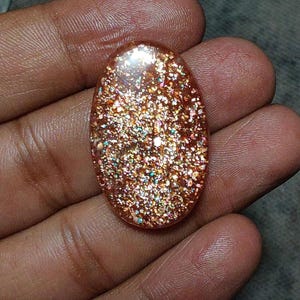 Può includere: Una pietra preziosa di pietra del sole di forma ovale con un colore di base caldo marrone rossastro e intense inclusioni metalliche riflettenti che creano un effetto scintillante e luccicante sull'intera superficie della pietra levigata.