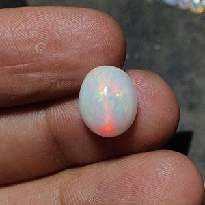 May include: A white oval opal gemstone with iridescent flashes of color. The opal is held between fingers, showcasing its polished surface and internal play of color, including red, green, and yellow hues. The gemstone is a natural, untreated opal.