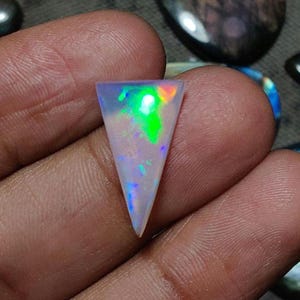 May include: A triangular opal gemstone with a pearly white base and iridescent flashes of green, blue, and yellow. The polished stone is held in a hand, showcasing its vibrant play of color. The opal's shape is a unique, elongated triangle.