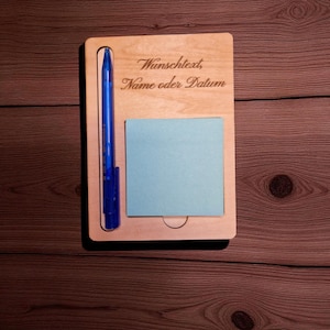 Könnte beinhalten: Ein Holz-Schreibtisch-Organizer mit einem blauen Stift und einem hellblauen Haftnotizblock. Der Text "Wunschtext, Name oder Datum" ist in das Holz graviert. Der Organizer befindet sich vor einem dunklen Holzmaserungshintergrund.
