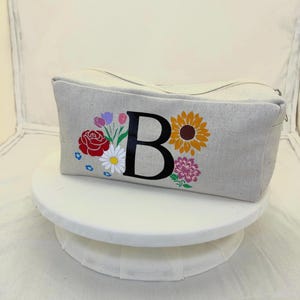 Puede incluir: Un neceser rectangular de lona gris claro con cierre de cremallera negro. La bolsa está decorada con una gran letra "B" negra rodeada de diseños florales coloridos, incluyendo una rosa roja, un girasol y otras flores.