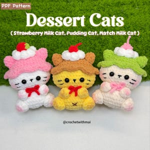 May include: Three crocheted cat plushies, each with a dessert-themed hat. The cats are white, yellow, and cream, with pink, brown, and green hats. The text "Dessert Cats" is at the top, with "Strawberry Milk Cat, Pudding Cat, Match Milk Cat" below. The artist's handle is @crochetwithmai.