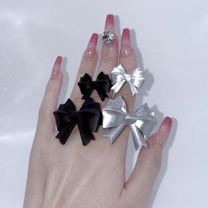 Peut inclure: Une main présente plusieurs bagues en forme de nœud papillon, noires et argentées. Une bague est ornée d'une grosse pierre précieuse transparente. Les ongles sont longs et peints avec un dégradé de rose et de rouge.