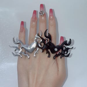 Puede incluir: Una mano muestra tres anillos abstractos: plateado, negro y plateado. Los anillos tienen un diseño fluido y orgánico. Un anillo transparente con una gran gema brillante está en el dedo medio. Las uñas son largas y están pintadas con un degradado de rosa y transparente.