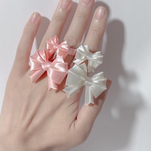 May include: A hand displaying four bow rings. Two rings are light pink, and two are white. The bows are made of a shiny, satin-like material. The rings are on a white background.