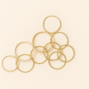 May include: A collection of gold-coloured rings, some with a smooth finish and others with a twisted rope design. The rings are arranged in an overlapping pattern against a neutral background.