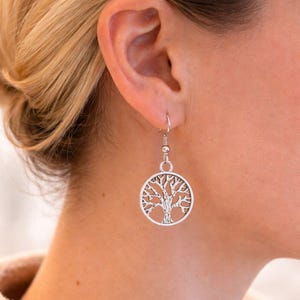 May include: Silver-coloured tree of life earrings. The earrings feature a circular pendant with a detailed tree design. The earrings are suspended from a simple hook. The image shows the earrings worn on a person.