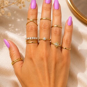 May include: A collection of ten gold-toned rings worn on fingers, featuring various designs including a crisscross band, a simple thin band, a row of small clear stones, a single solitaire stone, and a decorative leaf-shaped accent piece.