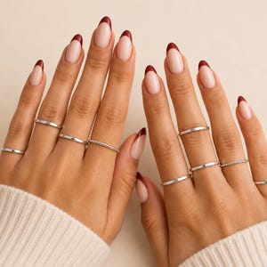 May include: A set of thin silver-toned rings worn on multiple fingers. The rings feature smooth bands and one twisted texture band. The fingernails are shaped in an almond style with a nude base and deep red tips, complemented by a white ribbed sleeve.