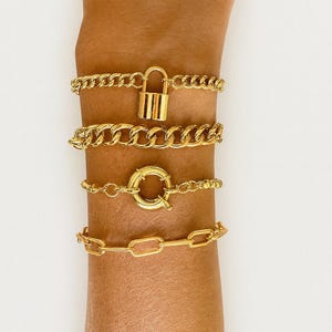 May include: A selection of gold-coloured bracelets. The bracelets feature different chain designs, including a curb chain with a padlock, a chunky curb chain, a chain with a circular clasp, and a link chain. The jewellery is shown on an arm.