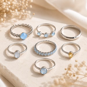 Könnte beinhalten: Ein Set aus sieben silberfarbenen Ringen mit verschiedenen Designs, darunter Bänder mit hellblauen runden Steinen, ein schlichtes poliertes Band, ein Ring mit einem einzelnen ovalen hellblauen Stein und ein Ring mit einem kleinen gelben Markisenschliff-Stein.