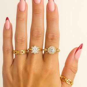 Puede incluir: Una mano muestra cuatro anillos de oro con diferentes diseños. Estos incluyen un anillo de cadena, un anillo floral con una piedra central y un anillo con una piedra grande, clara y redonda. Las uñas están pintadas con una base rosa y una punta roja.