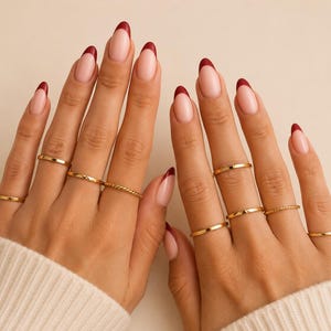 May include: Hands displaying gold rings and a French manicure with a burgundy tip. The rings are different styles, including a twisted band and simple bands. The nails have a light pink base.