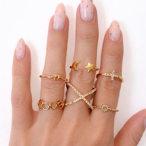 May include: A hand adorned with various gold rings. The collection includes a knot ring, a ring spelling "love", a moon and star ring, a cross ring, and a ring with a small gemstone. The hand has light pink nails.