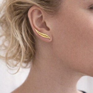 Könnte beinhalten: Ein goldfarbener Ohrschmuck in Form eines Blattes wird am Ohr getragen. Der Schmuck hat ein detailliertes, strukturiertes Design. Der Ohrschmuck wird am oberen Ohr getragen, und die Person trägt ein schwarzes Oberteil.