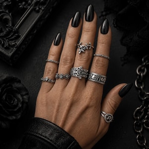 May include: A set of eight silver-toned rings worn on fingers, featuring various designs including a hamsa hand, a leaf motif, geometric patterns, and thin bands with intricate engravings, all displayed on a hand with black fingernails.
