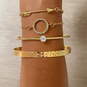 May include: A selection of gold bracelets. The bracelets feature an arrow design, a circle of small clear stones, a thin band with a single clear stone, and a hammered metal bangle. The bracelets are stacked on a wrist.