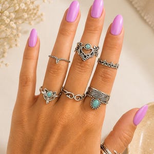 May include: A set of seven silver-toned rings worn on fingers, featuring various designs including filigree patterns, twisted bands, and three rings accented with small, oval-shaped turquoise-coloured stones set in metal mountings.