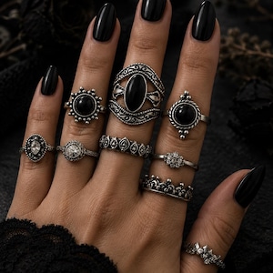 May include: An assortment of silver rings featuring black gemstones and intricate patterns. The rings, including a crown design, are worn on a hand with black nail polish, highlighting the jewellery's vintage and gothic style.